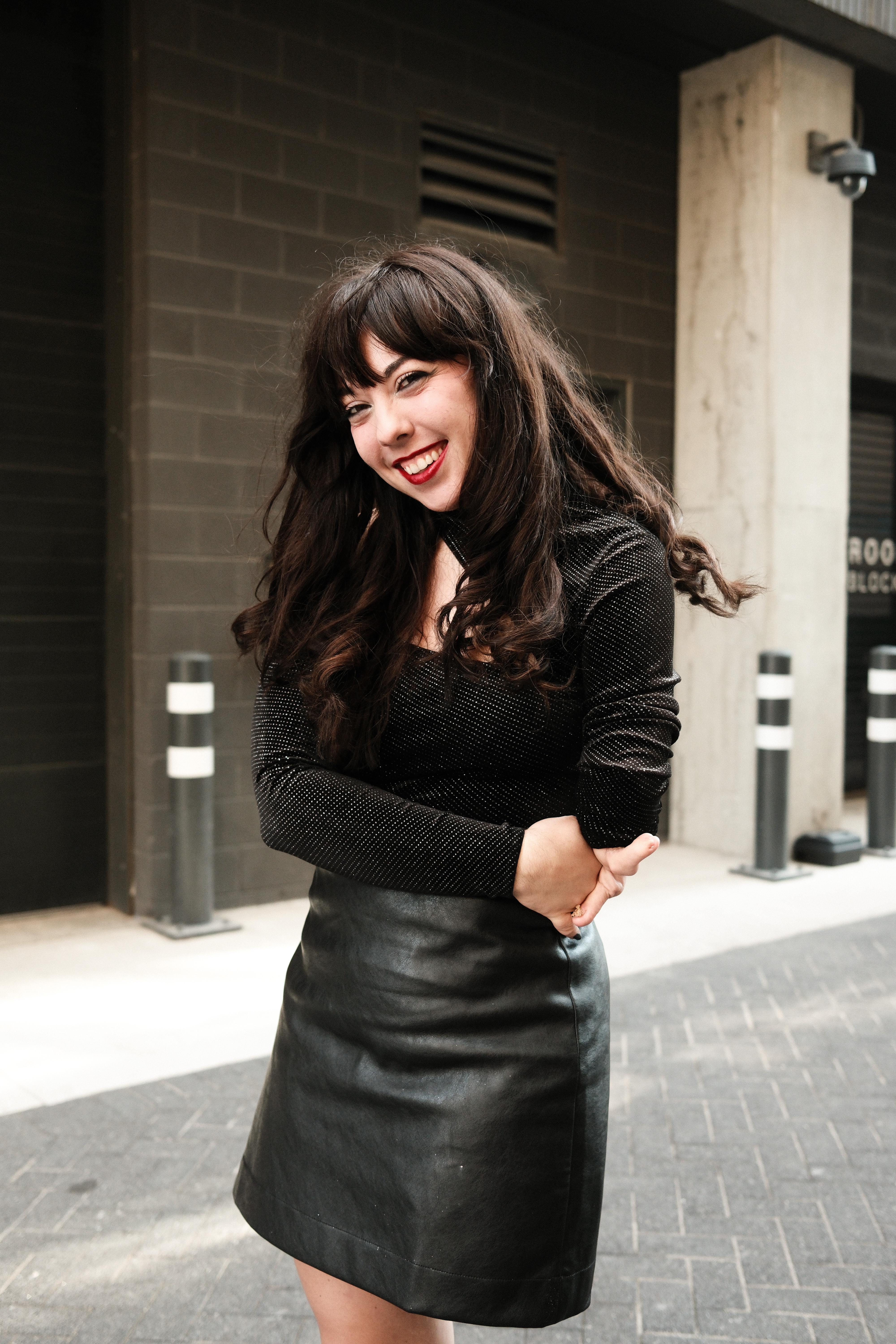 Jessica Jordan Ping, a white woman with long black hair stands in an alleyway in Chicago, IL wearing a long sleeve black sparkle top and a black leather mini skirt standing on her one leg and her arms crossed in front of her, smiling wearing red lipstick