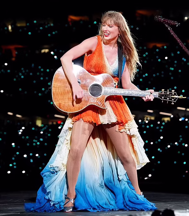 Taylor swift on stage during the acoustic set of her eras tour show wearing an orange, blue, and white roberto cavalli dress